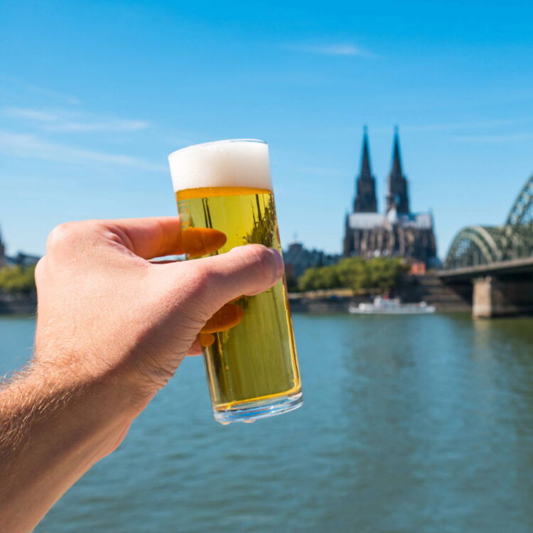 Eine Hand hält ein Glas Kölsch vor die Kamera. Im Hintergrund sind der Rhein und der Kölner Dom zu sehen.